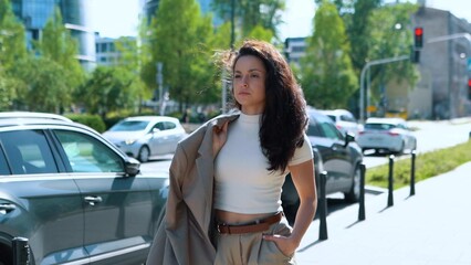 Fototapeta na wymiar Portrait of concentrated serious Caucasian woman carrying jacket on shoulder walking outside on sunny day. Young confident business lady on street in big city. Successful business people concept.