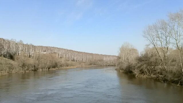 Tall bare trees at a bend in the bed of a calm small river flowing through a hilly valley on a clear spring evening. Inya River, Novosibirsk, Siberia, Russia.