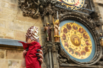 Female hand is holding traditional czech cookie trdelnik on background of Astronomical clock in Old Town of Prague city.