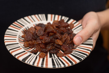 Close-up shot of a person's hands holding a small striped plate filled with juicy, dark raisins.