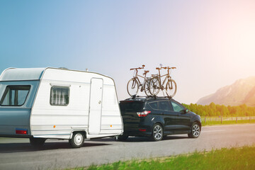Black suv with bicycles mounted on top tows a white caravan on a road trip with scenic mountains in...