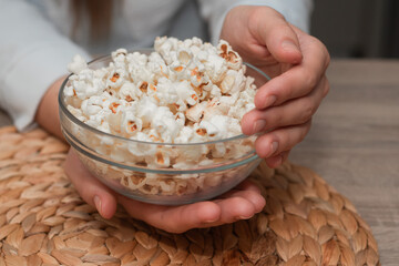 Woman Holding Bowl of Fresh Popcorn. Close-up of a woman's hands holding a transparent bowl filled with freshly popped popcorn, on a natural woven mat.