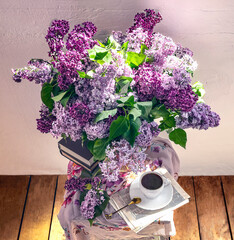 A beautiful bouquet of purple lilac flowers in a crystal vase and a white vintage cup of coffee, books on a light background. Spring still life in sunlight