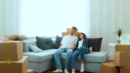 Fototapeta na wymiar Caucasian joyful boyfriend and girlfriend moving in together in new house. Tired happy young married couple sitting down on sofa in room with many boxes on moving day. Home concept
