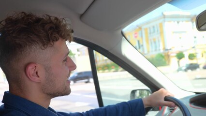 Close up shot of happy Caucasian man driving a car and smiling. Cheerful positive guy with curly hair drives his auto looking at road. Side view. Young male driver inside the vehicle