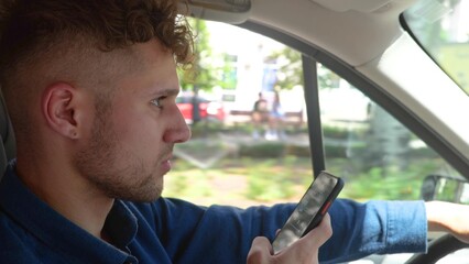 Young handsome serious man driving a car and typing on smartphone while looking at road. Side view. Close up of male driver using mobile phone while driving a vehicle. Taxi cab