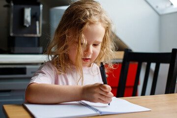 Close up photo of a blonde little girl drawing something in her notebook sitting at the table in the kitchen