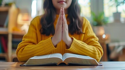 Woman in yellow sweater sitting, hands folded in prayer over open bible at table