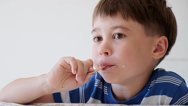 Handsome boy eating popsicle on a stick. He bites off the ice cream with pleasure. Close-up. Child eat in the kitchen or at the dining room table. Tasty dessert. Slow motion. High quality 4k footage