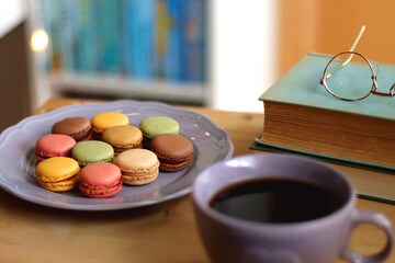 Purple plate filled with pastel macarons, cup of tea or coffee, vintage books and reading glasses on the table. Colorful bookcase in the background. Selective focus.