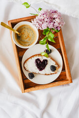 Süsses Frühstück mit einer Kaffeetasse und eine Scheibe Brot mit Konfitüre in Herzform. Braunes Serviertablett, weiße Bettdecke, Draufsicht.