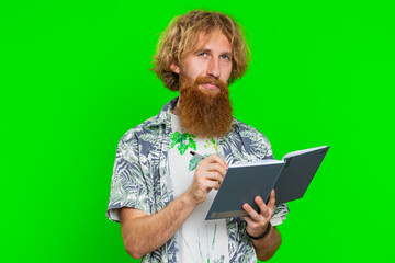 Thoughtful journalist young man making notes, writing down thoughts with pen into notepad notebook diary, to do list, good idea. Handsome redhead student guy isolated on green chroma key background