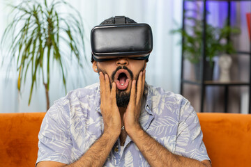 Indian man using virtual reality futuristic technology VR app headset helmet to play simulation 3D 360 online video game, watching film movie at modern home apartment. Guy in goggles sitting on sofa