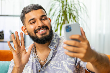 Happy Indian man blogger taking selfie on smartphone, communicating video call online with subscribers, recording stories for social media. Guy at modern home apartment living room sitting on sofa