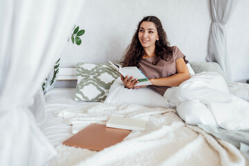 An attractive young woman is lying in bed on pillows and reading a book. A smiling curly-haired...