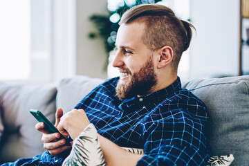 Cheerful hipster guy enjoying internet connection at home for chatting in social networks via...