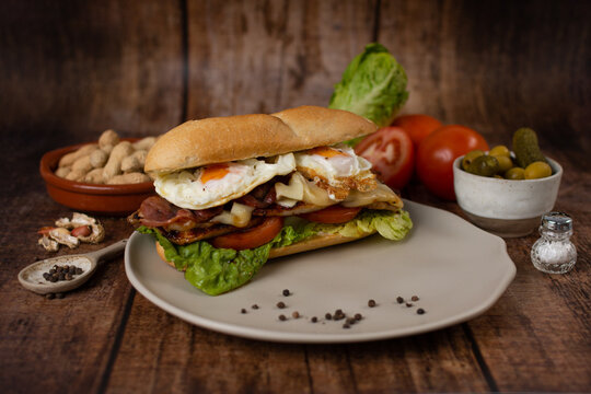 A chivito sandwich, which includes roast pork loin, bacon, cheese, lettuce, tomato, fried egg and mayonnaise, served on a toasted bun accompanied by olives and peanuts