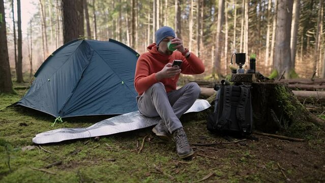 Morning at campground in fall forest. Tursit is browsing Internet on his smartphone while sitting on hiking mat near tent and drinking a beverage from a cup. Travel, people and healthy lifestyle. 