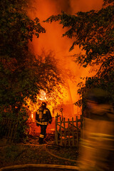 Firefighters silhouette against background of orange glow from fire in country house at night. Fire...