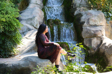 Latin woman, brunette with curly hair, young and beautiful, sitting on the rock next to a waterfall among the vegetation. The woman is serious and pensive with her gaze fixed on infinity.