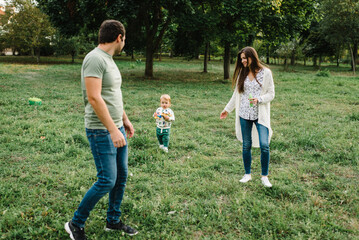 Fototapeta premium Happy mother, father hug baby son walking in garden at sunset. Family spending time together outdoors. Child with parents happiness playing in green grass in park. Children's day. Friendly family.