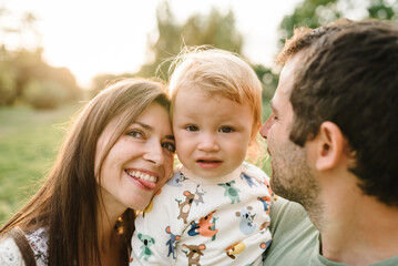 Parents hugging kid and happiness playing in green grass in park. Happy mother, father hug baby son walking in garden at sunset. Family spending time together outdoors. Children's day. Friendly family
