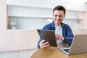 An enthusiastic young professional in a contemporary kitchen setting uses both a tablet and a laptop, displaying a cheerful and engaged demeanor.