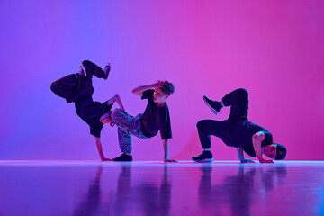 Group of teenagers, boys in casual attire dancing breakdance in mixed neo n light against vibrant...