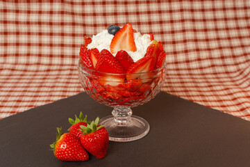 Strawberries with cream are served in an elegant glass cup on a slate base. The background is adorned with a red checkered tablecloth