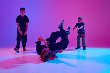 Three young dancers in casual attire perform a vibrant breakdance routine in mixed neon light against vibrant gradient background. Concept of sport and hobby, music, fashion and art, movement. Ad