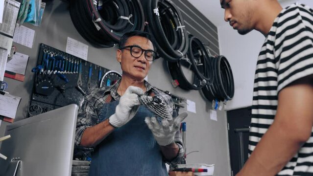 Shopkeeper senior asian man introduce and tell detail of gear to customer at tool and equipment bike shop. Small business and dealer concept