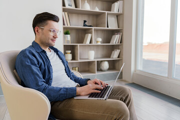 A focused young professional works on his laptop while seated in a stylish armchair in a serene and elegant living space, demonstrating concentration and comfort.