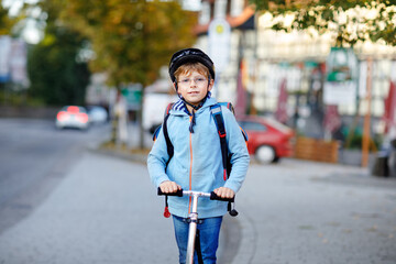 Active school kid boy in safety helmet riding with his scooter in the city with backpack on sunny day. Happy child in colorful clothes biking on way to school.