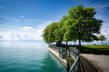Blick entlang des Zauns einer Seeuferpromenade mit Aussicht auf den See und dicht belaubten Bäumen, sowie Sitzbänken auf einer Wiese am Uferrand