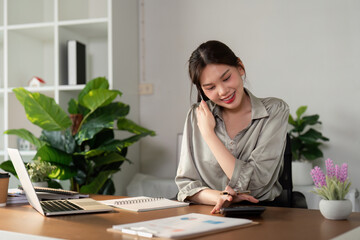 Happy smiling asian business woman working on laptop at office, using smart phone. Businesswoman sitting at office