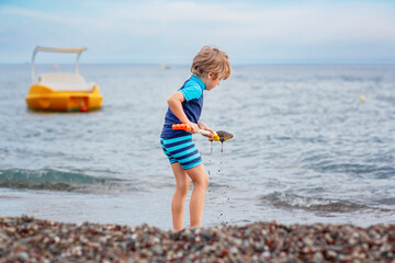 Adorable little blond kid boy building pebble stone castle on beach. Funny child playing with...