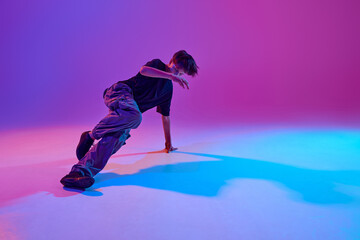 Stylish dressed junior dancer, guy performing breakdance in motion in mixed neon light against...