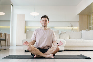 A man sits cross-legged on a yoga mat, meditating peacefully in a lotus position within a bright and modern living space, embodying tranquility and mindfulness.