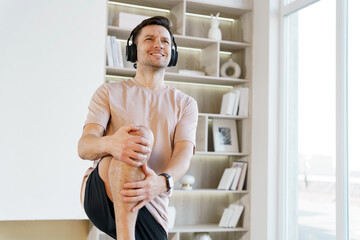 A smiling man wearing headphones holds a knee stretch in a sunny, book-lined room, enjoying a relaxing fitness session.