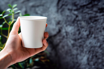 Hand holding a white disposable cup on a blurred grey background. Blank space for product placement or advertising text