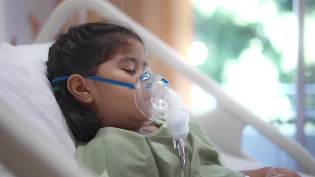 A girl is hooked up to an oxygen tank in a hospital bed. She is wearing a green gown and she is in pain.