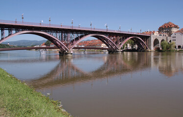 Photo of the iron arch bridge reflected in the Drava River and historical buildings on the embankment in the city of Maribor, Slovenia