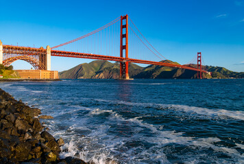Panoramic view of famous Golden Gate bridge seen from Crissy Field, San Francisco (USA) on a clear sunny morning with blue sky. Iconic red silhouette with waves on the stony shore in the foreground.