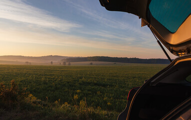 Rear of car with open automobile trunk with spring Czech landscape in misty fog at sunrise. Trip,...