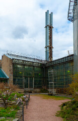 Aged urban greenhouse with large glass panels, set against a modern city tower, located within a lush city park setting