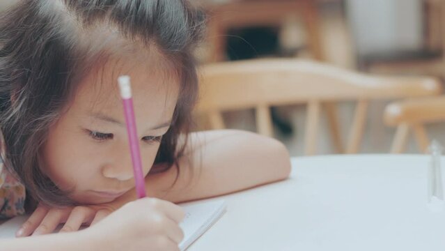 Face and eyes close up of adorable Asian kid girl who is writing with determination and concentrating on notebook shows concept of education and studying of child in classroom for school student.