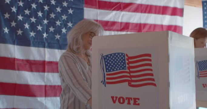 Wide shot of mature woman voter smiling  and voting behind a polling booth with an American flag in the background