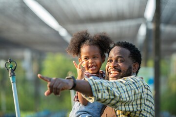 Happy african black parents dad father with daughter child on piggyback teasing fun in garden greenhouse. Black daughter kiss cheek dad and neck riding in vegetable greenhouse garden in light sunset - Powered by Adobe