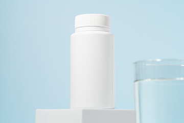 White mockup of a jar of pills on a pedestal and a glass of water on a blue background. Concept of pharmacy, medicine, vitamins, dietary supplements, and disease treatment.