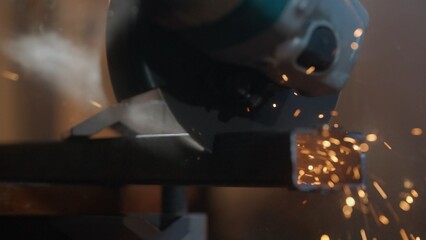 Craftsman working with grinder at industrial plant, man grinding iron detail, sparks flying around and falling on the ground in dark workshop. Close up shot.